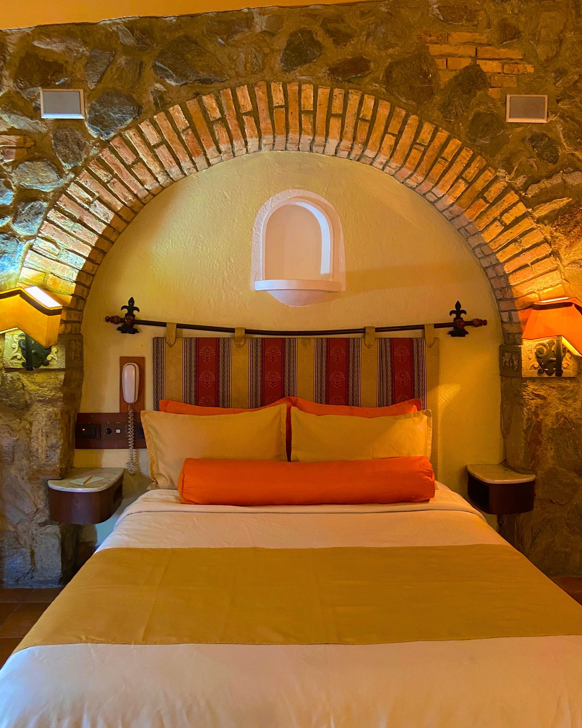 Mexicana Premium: cama bajo un arco de ladrillo y piedra, con cabecero de tela típica y cojines naranjas